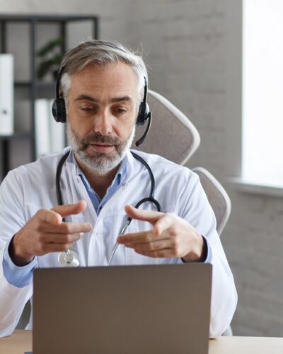 Portrait of senior grey-haired male doctor in his office using laptop for video chat with a patient. Online consultation with doctor for diagnoses and treatment recommendation. Telehealth concept.