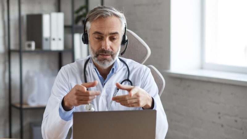 Portrait of senior grey-haired male doctor in his office using laptop for video chat with a patient. Online consultation with doctor for diagnoses and treatment recommendation. Telehealth concept.